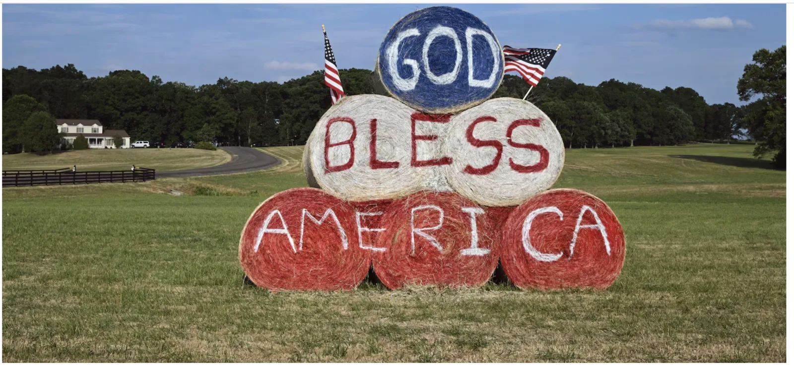 Round hay bales painted red, white and blue spelling GOD BLESS AMERICA flanked by two American flags in a rural field